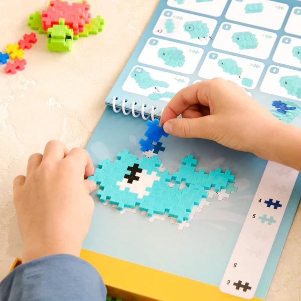 Plus Plus Activity Pad | Ocean showing hands assembling a blue whale with numbered guides and pieces on a flippable pad.