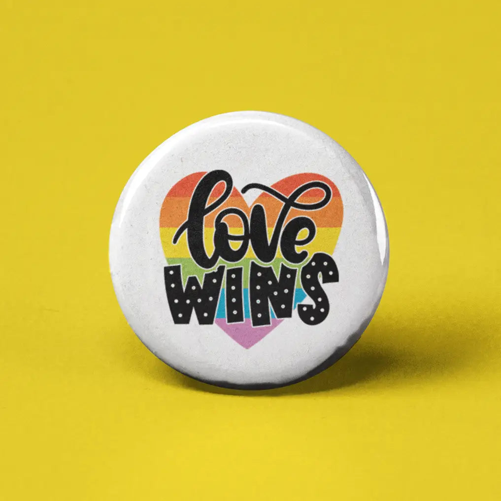 Button | Love Wins with rainbow heart background and bold black text on white 1.25-inch button on yellow surface