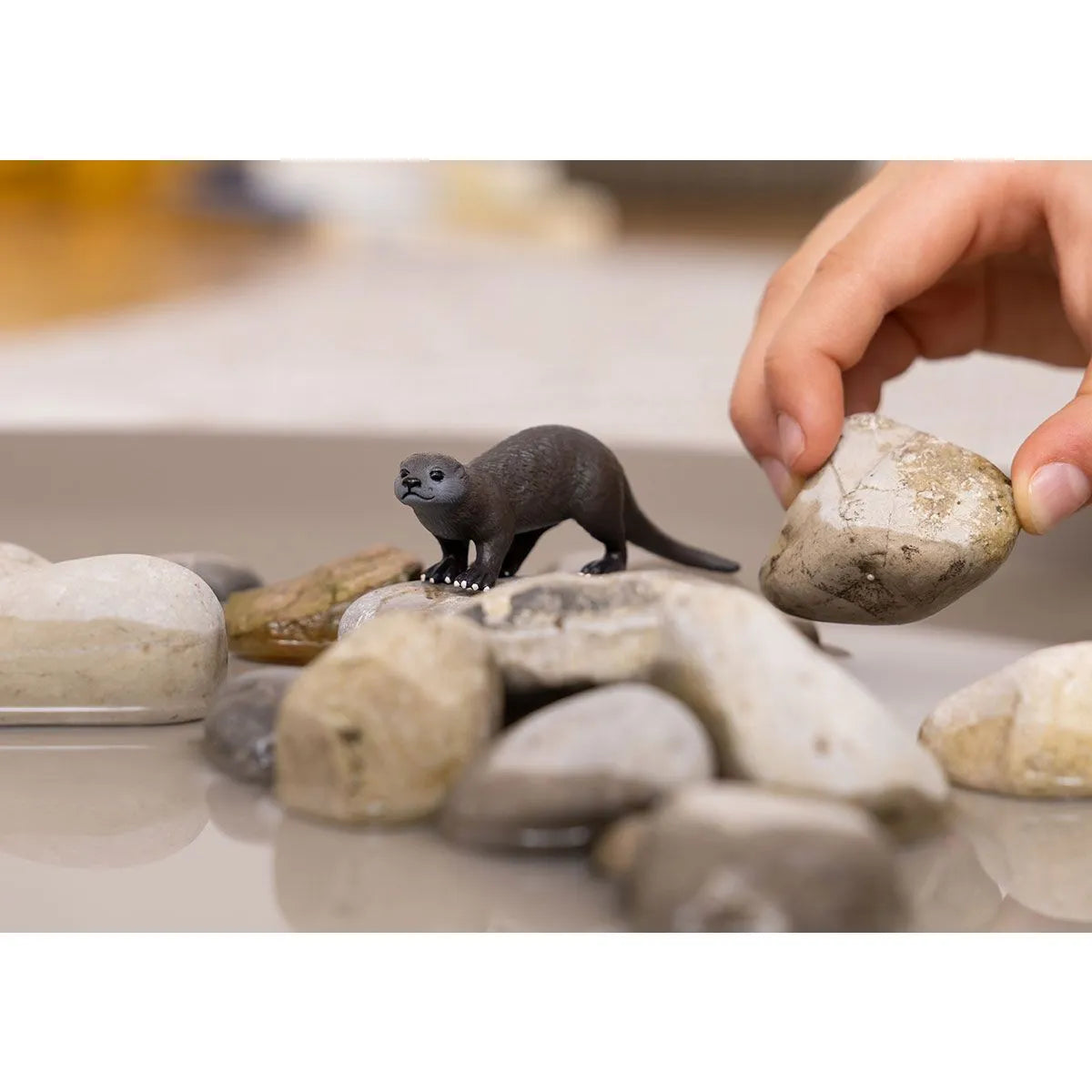 Otter figurine among smooth stones with a hand placing a rock nearby, showing playful nature Otter toy.