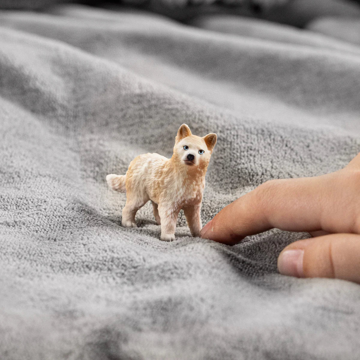 Small cream-colored Arctic Wolf Cub figurine with bright blue eyes beside a human finger on soft fabric.