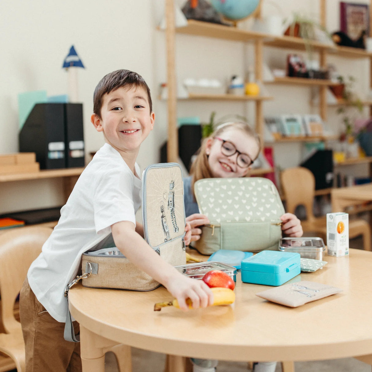 Two children opening insulated lunch boxes on a table with snacks and drinks, Lunch Box | Robot Playdate.