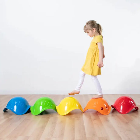 Child in yellow dress balancing on colorful Bilibo toys arranged in a row on wooden floor for imaginative play.