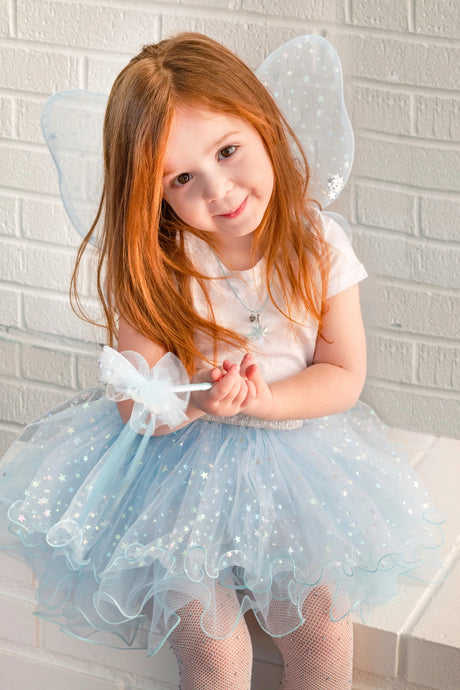 Young girl wearing light blue fairy wings and tutu with silver stars holding a matching star wand sitting by white brick wall.