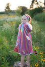 Young girl outdoors wearing Rainbow Princess Cape with fluffy feather trim and shimmering rainbow colors.