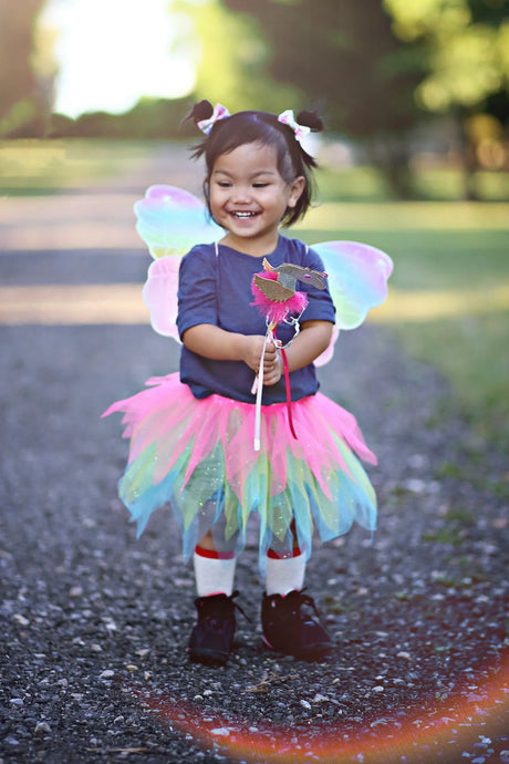 Happy toddler wearing Neon Rainbow Skirt Wings & Wand Set with colorful tutu, glitter wings, and matching wand outdoors.