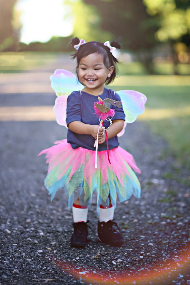 Happy toddler wearing Neon Rainbow Skirt Wings & Wand Set with colorful tutu, glitter wings, and matching wand outdoors.