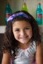 Smiling girl wearing a Midnight Metallic Headband with a shiny purple and blue bow, perfect for ages 3 and up.