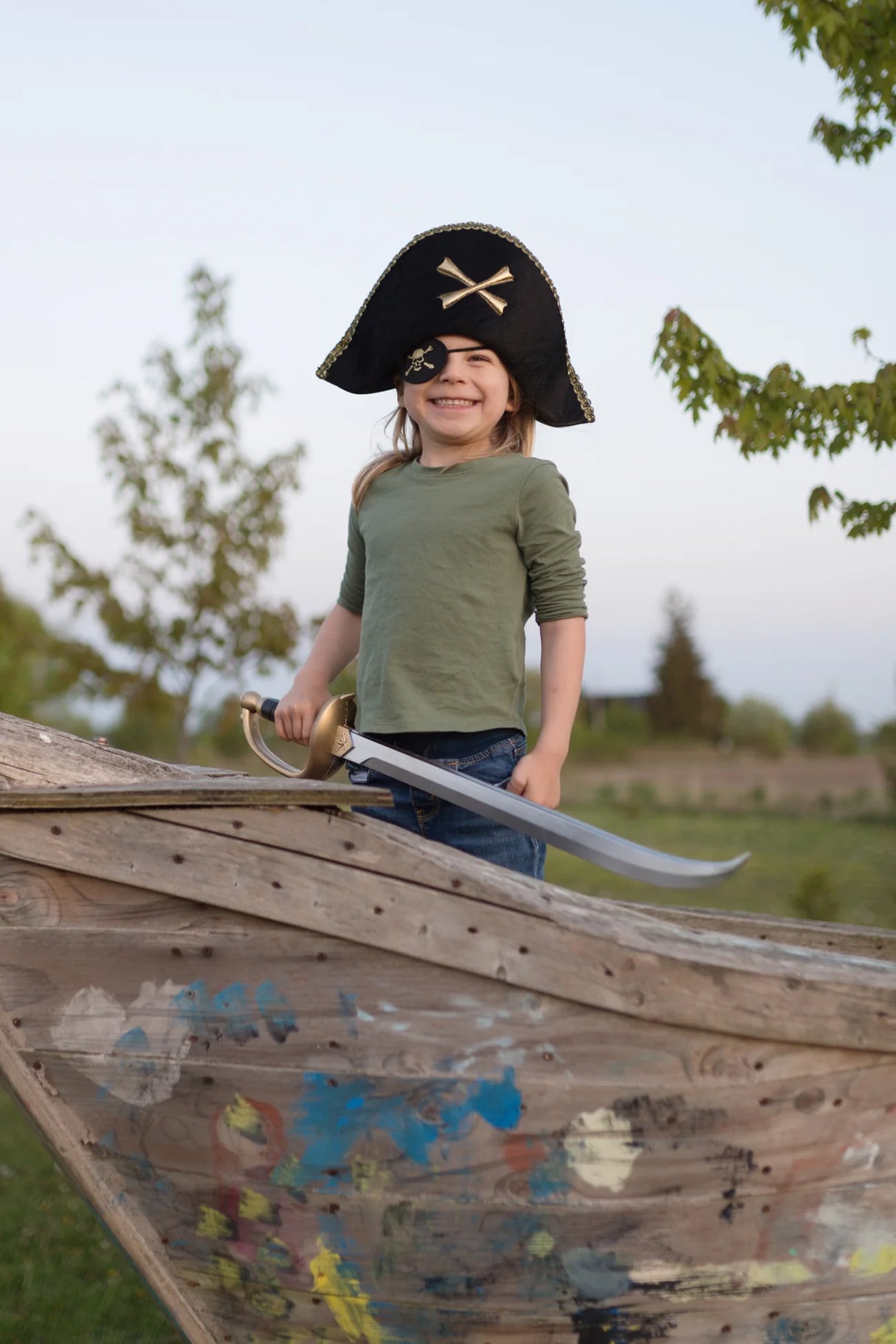 Child wearing a Pirate Captain Hat with gold trim and crossbones, smiling on a wooden play ship outdoors.