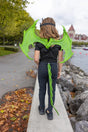 Child wearing bright Green Dragon Wings and tail accessory walking outdoors near water and autumn leaves.