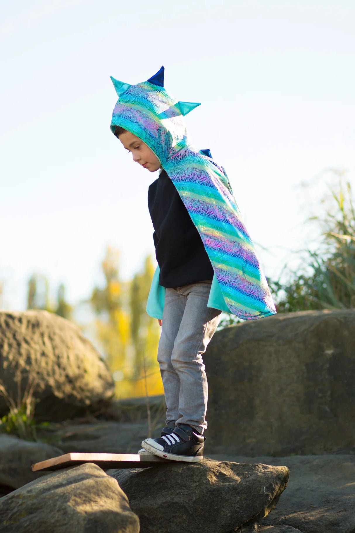 Child wearing Unicorn Dragon Reversible Cape with shiny dragon scales hood outdoors on rocks balanced on plank