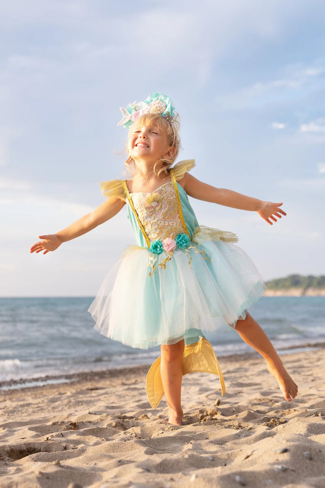 Young girl joyfully wearing the Mermalicious Dress with Tail at the beach, featuring a shimmery sea blue skirt and floral details.