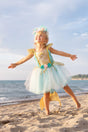 Young girl joyfully wearing the Mermalicious Dress with Tail at the beach, featuring a shimmery sea blue skirt and floral details.