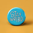 Button | Stay Weird round blue button with white playful text on a yellow background measuring 1.25 inches in diameter