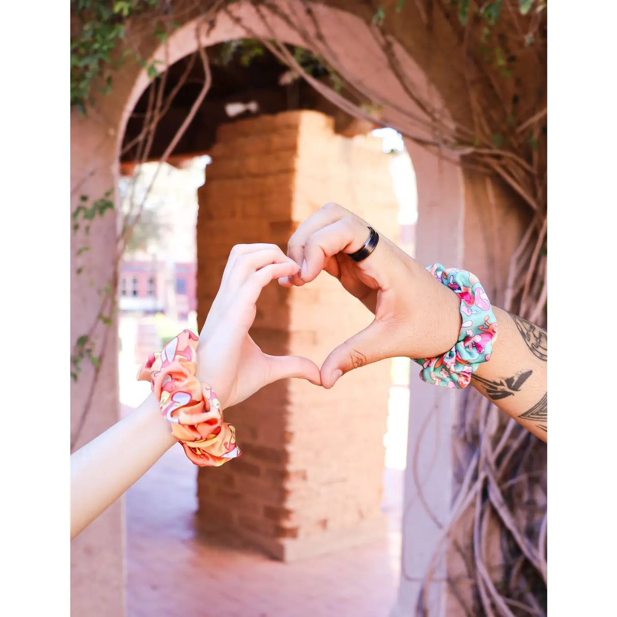 Two hands forming a heart shape wearing colorful Axolotl Hair Scrunchie satin fabric accessories outdoors.