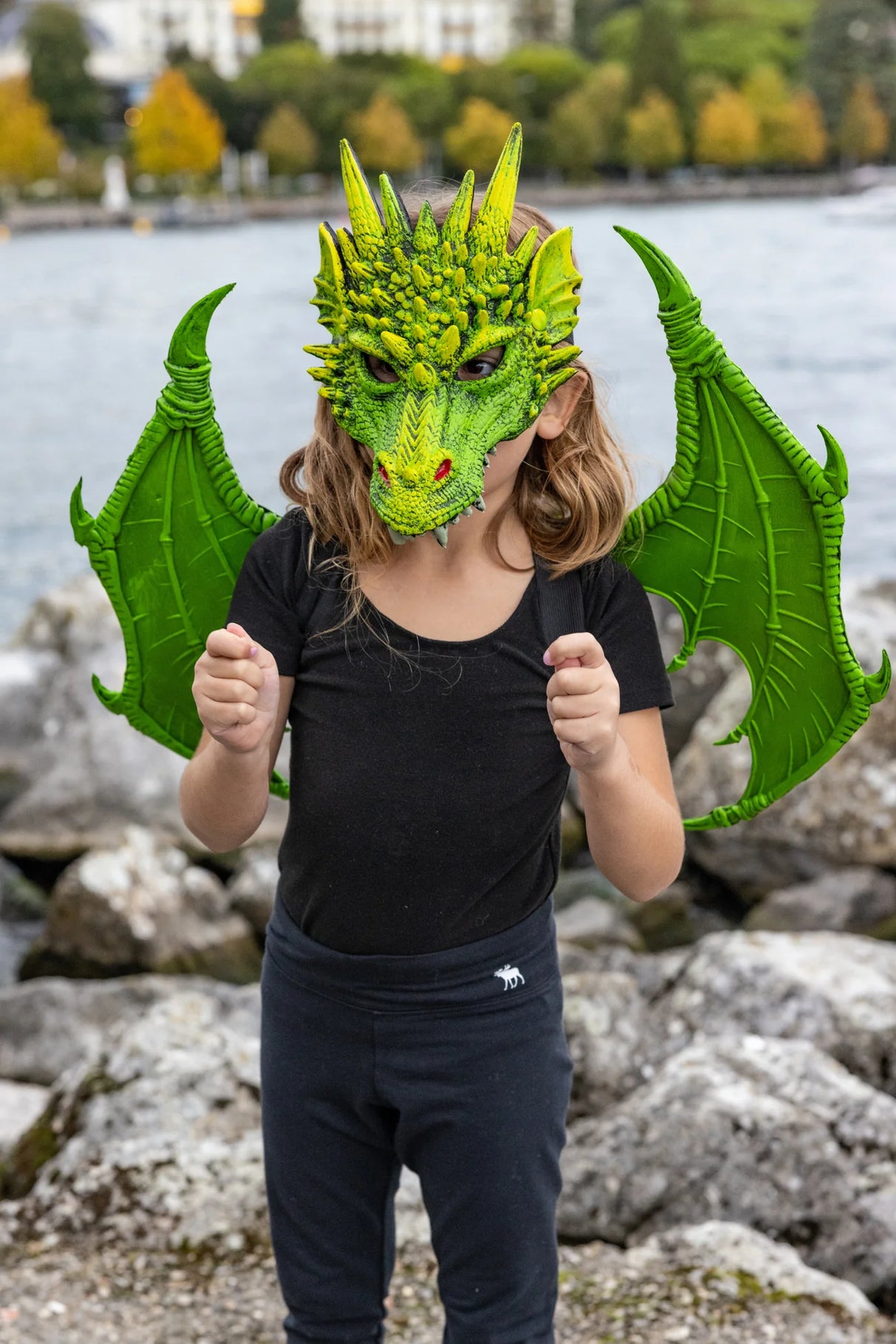 Child wearing a green dragon mask with Green Dragon Wings outdoors near water and rocks, mask sold separately