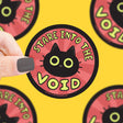 Hand holding Stare Into the Void Sticker featuring a black cat on red background, durable vinyl, weatherproof design.
