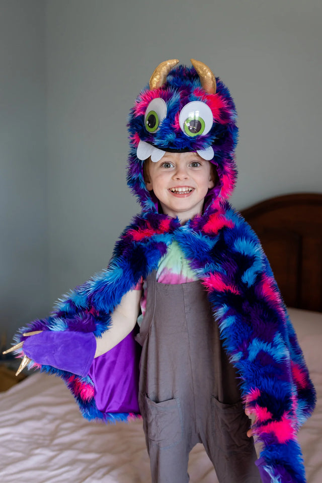 Child wearing Goober the Monster Cape featuring bright blue, pink, and purple fur with googly eyes and gold horns on the hood.