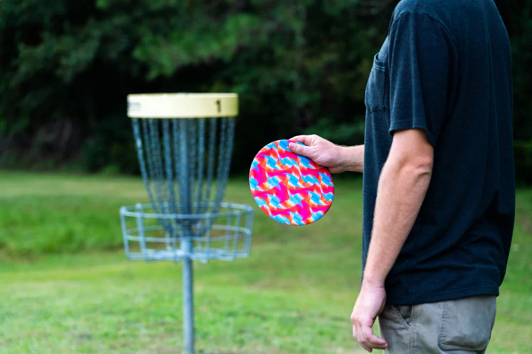 Person holding a colorful Waboba Wingman Disc PRO ready to throw towards a disc golf basket outdoors.
