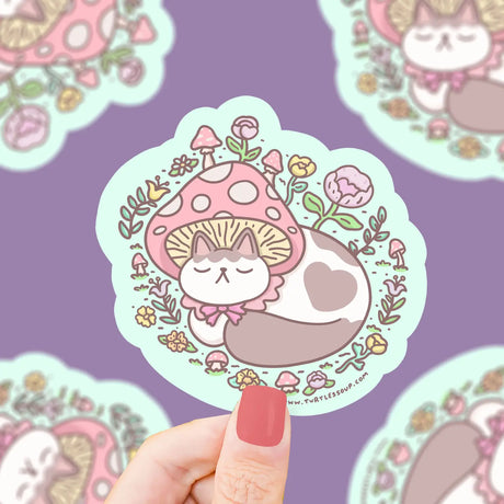 Hand holding Mushroom Cap Kitty Sticker featuring a cute cat sleeping under a pink mushroom cap with floral accents.