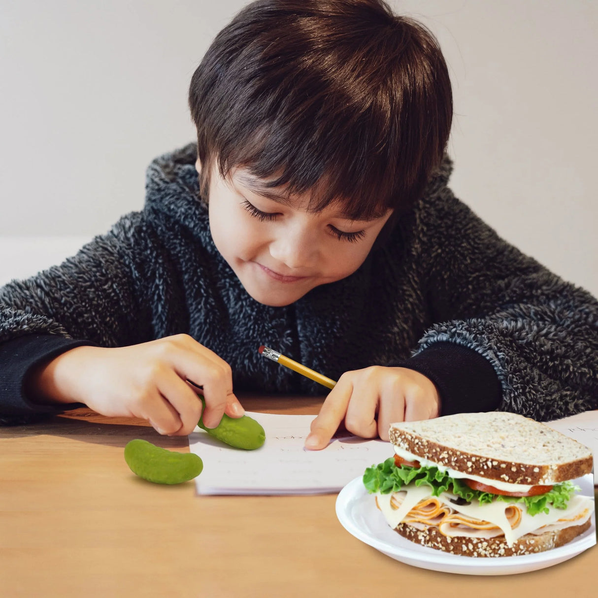Child using Pickle Erasers shaped like mini pickles to correct mistakes while writing at a table with a sandwich nearby.