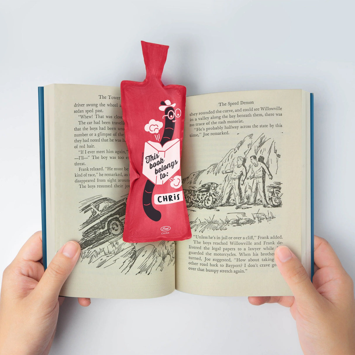 Bookie Cushion Bookmark in red, placed inside an open book to hold a reading spot with a fun design.