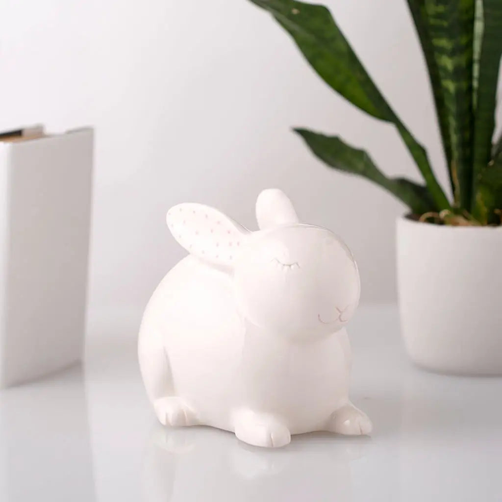 Ceramic Bunny Money Bank in white with pink ears, sitting on a white surface near a green plant and books.