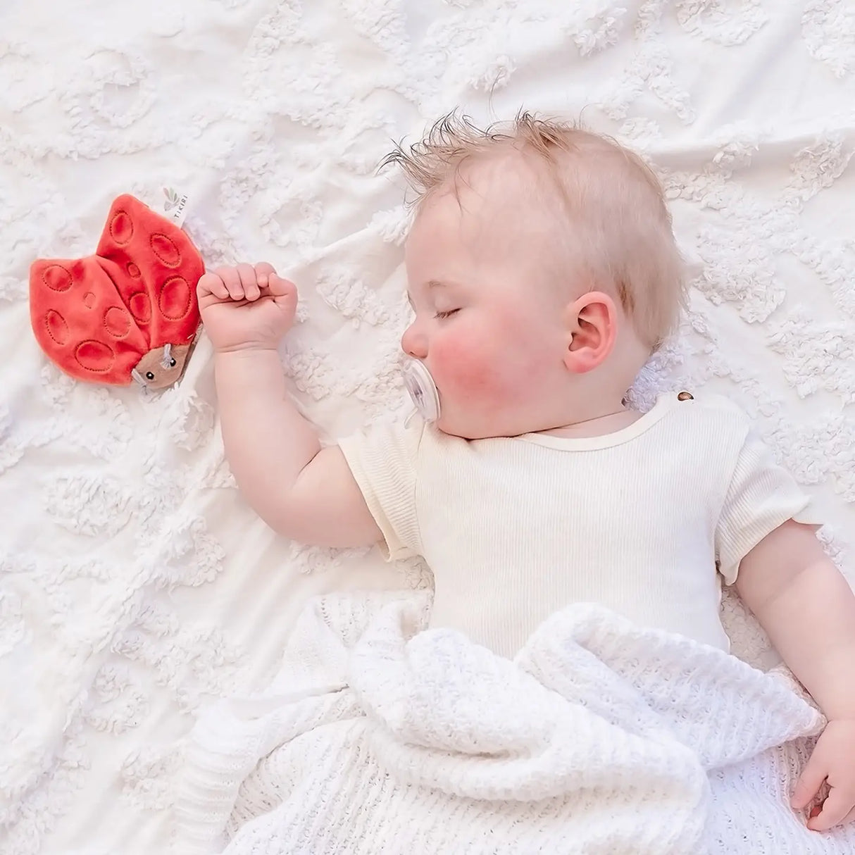 Sleeping baby next to a red and brown ladybug toy made of Organic Crinkle Fabric | Ladybug on white blanket.