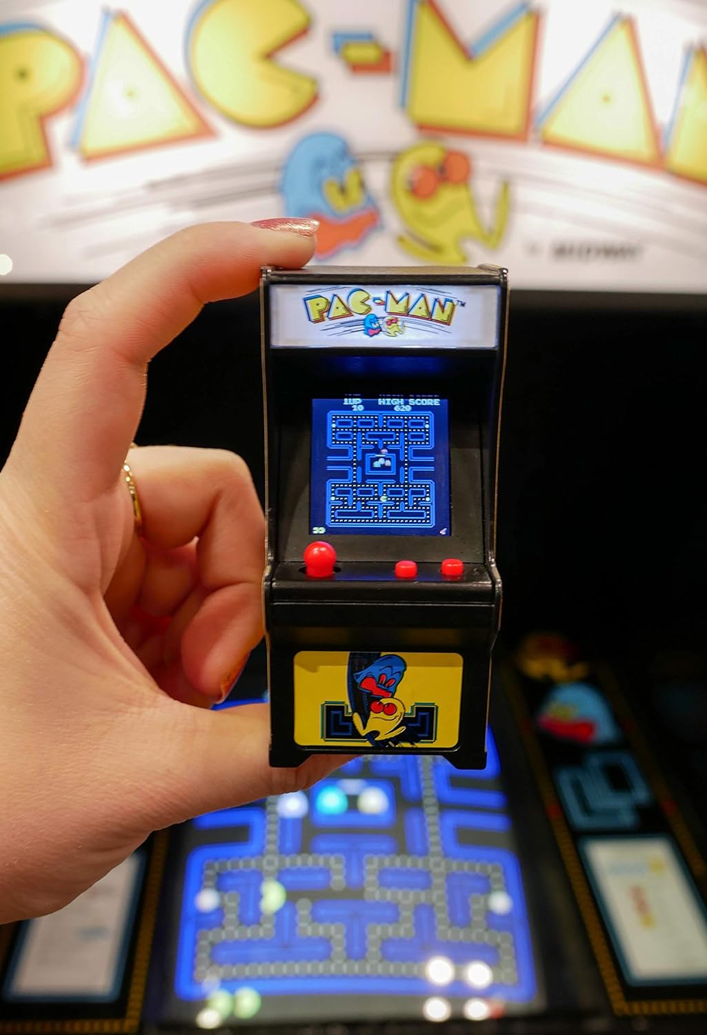 Hand holding Tiny Arcade Pacman with screen showing gameplay and colorful ghosts in background.