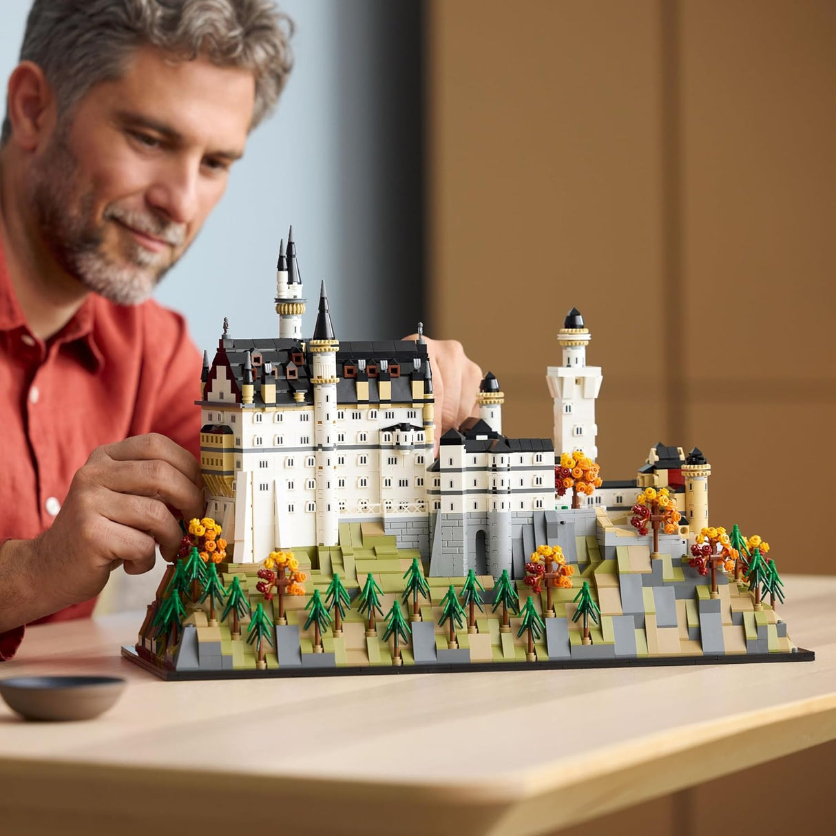 Adult building a detailed LEGO model of Neuschwanstein Castle with customizable fall foliage on a wooden table.