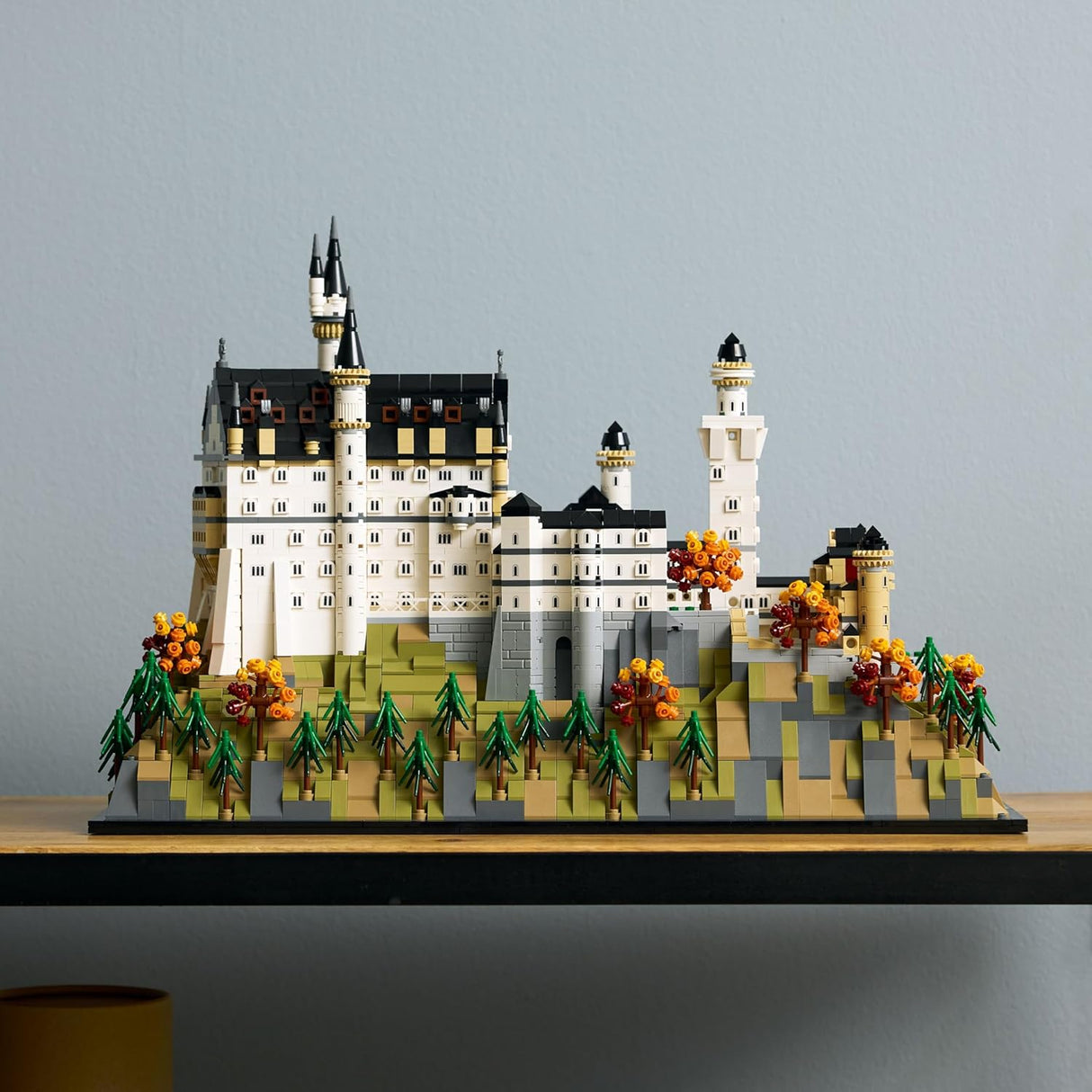 Detailed model of Neuschwanstein Castle with autumn trees and green pine trees on a wooden shelf against a gray wall