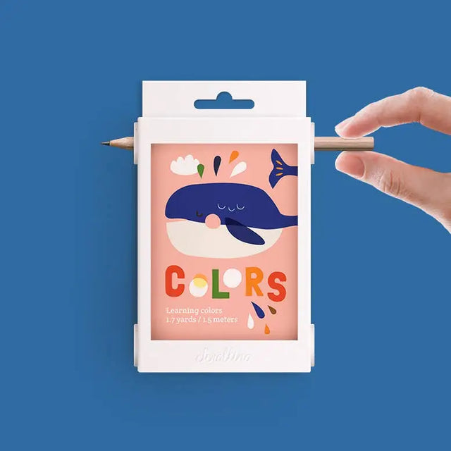 Scrollino Colors Learning Book with colorful whale illustration held by hand on blue background.