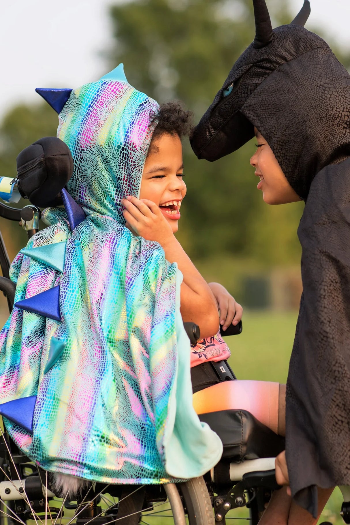 Two children play outdoors wearing the Unicorn Dragon Reversible Cape with shimmering rainbow and black dragon sides.