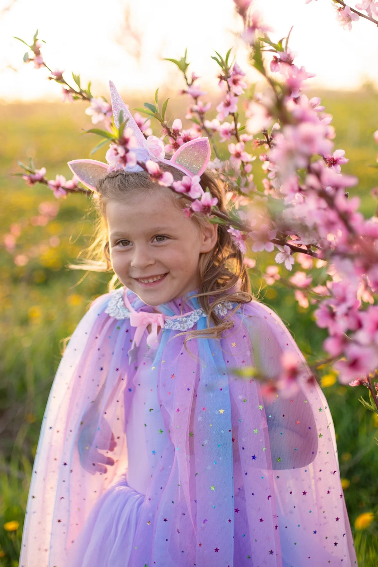Young girl wearing Rainbow Unicorn Cape & Headband with sparkly pastel cape and unicorn headband outdoors among pink blossoms