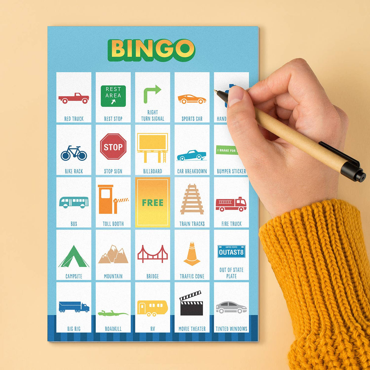 Hand holding pen marking spaces on colorful travel bingo game pad with various road trip icons