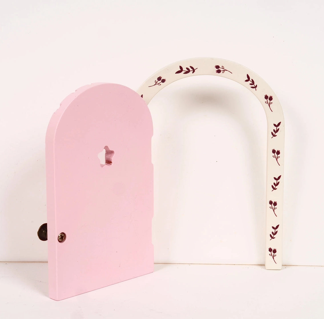 Small pink wooden fairy door with decorative white arch featuring red floral designs, perfect for magical play spaces.