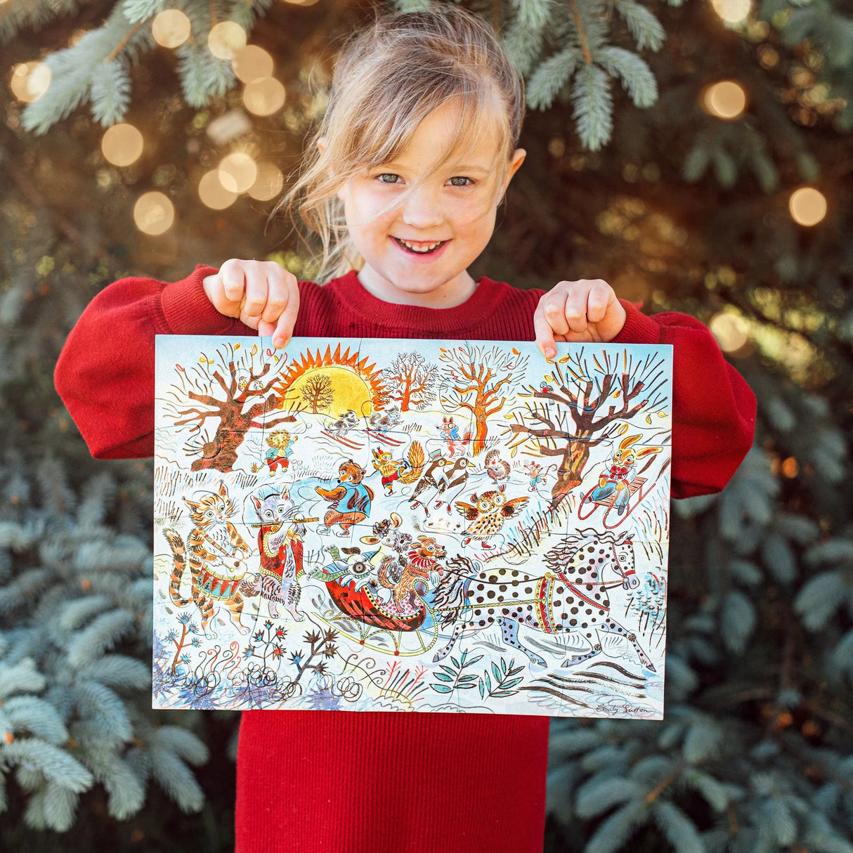 Child in red sweater happily holding completed vibrant winter-themed animal puzzle outdoors by tree lights