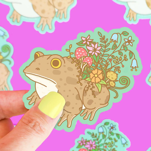 Hand holding Botanical Toad Planter Sticker showing a toad with colorful plants and mushrooms growing on its back.