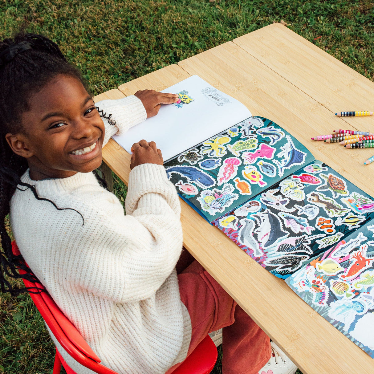 Girl smiling while using colorful stickers in Learn to Draw | Under the Sea drawing book outdoors on wooden table