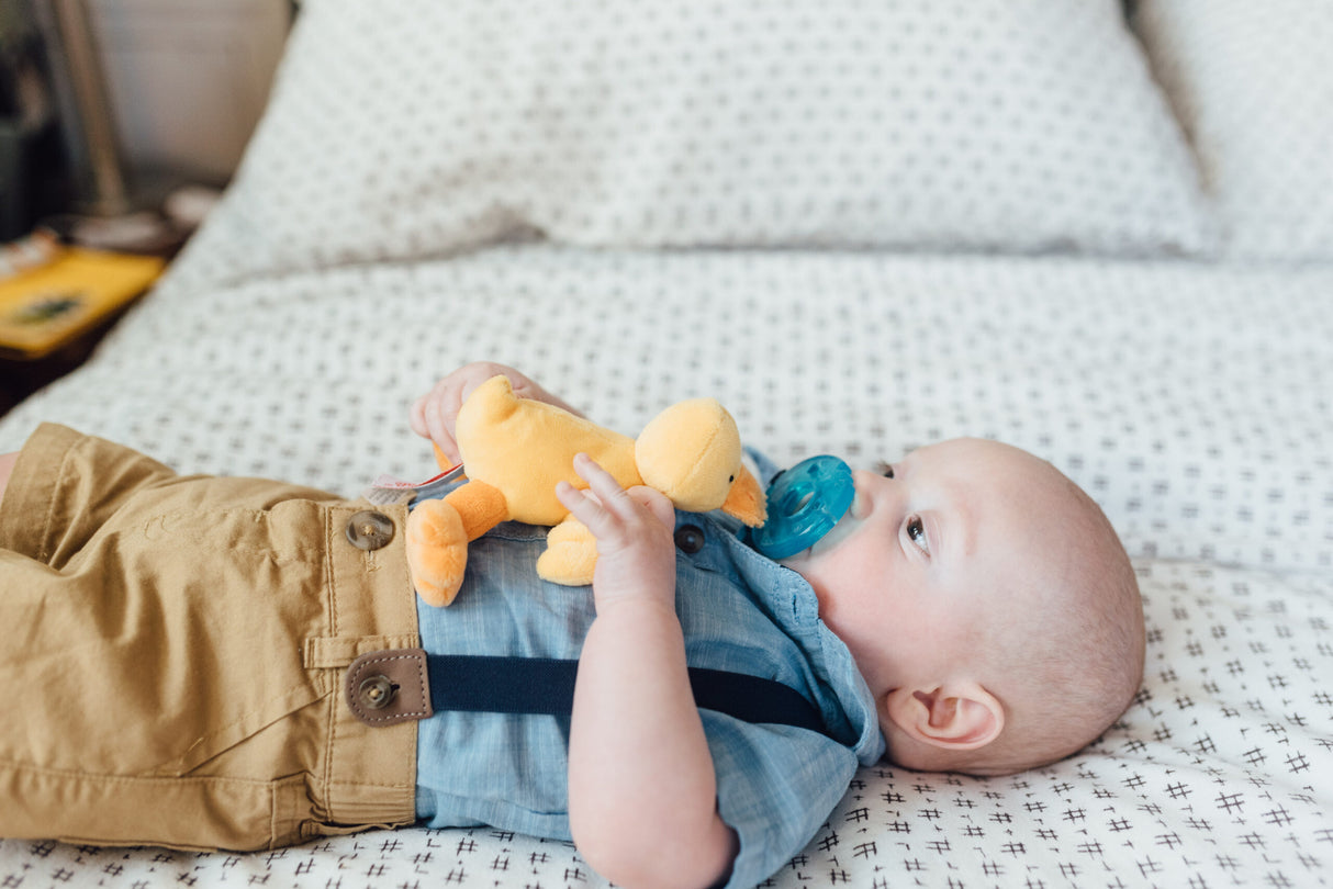 Baby lying on bed holding WubbaNub | Duck pacifier with yellow plush duck attached for soothing comfort.