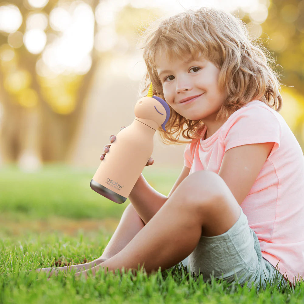 Child sitting on grass holding an Asobu Water Bottle | Unicorn with a soft unicorn head lid outdoors.