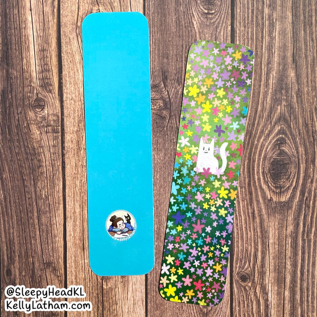 Cat & Flowers Bookmark showing a white cat among colorful flowers on one side and solid blue with artist logo on the back.
