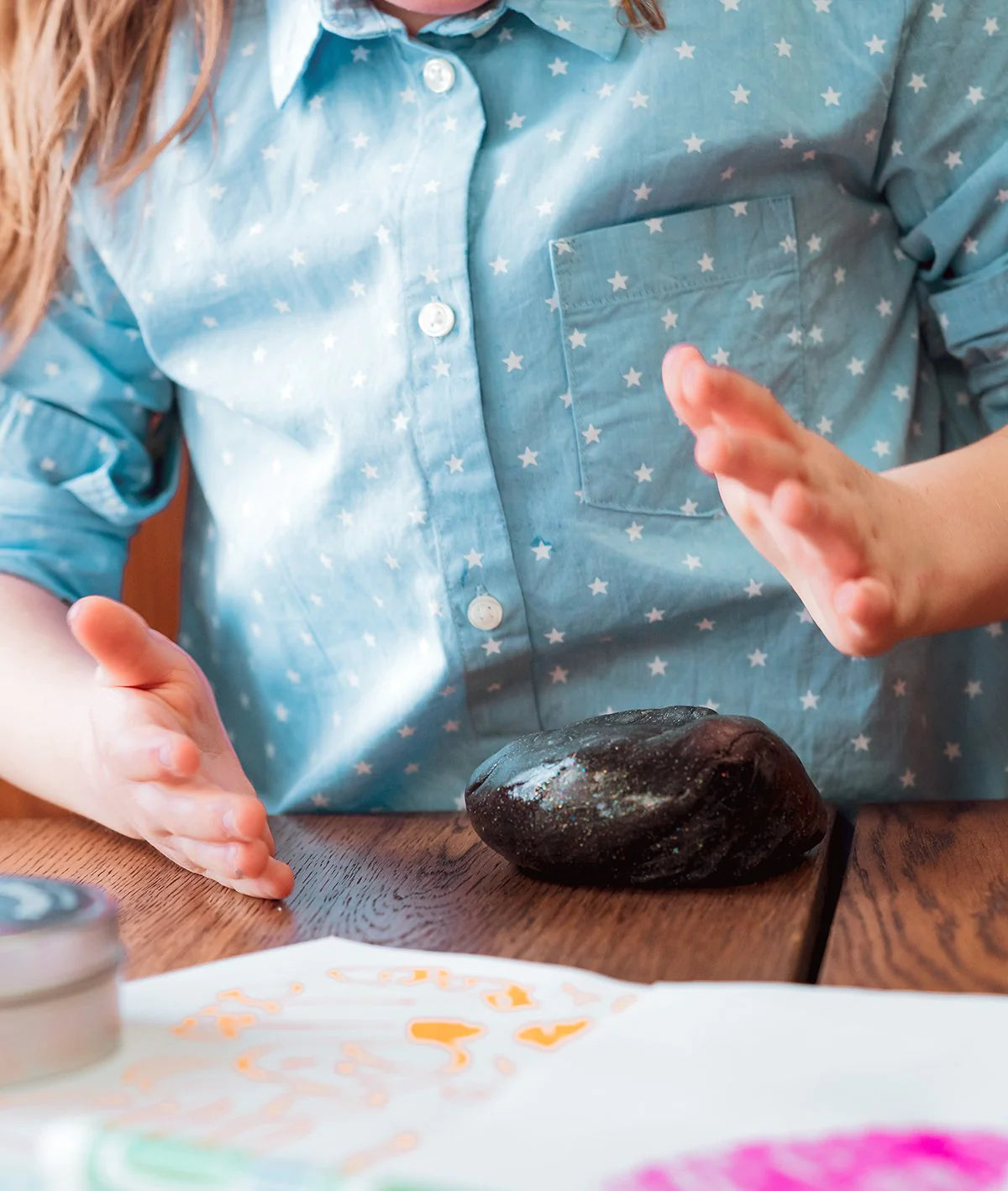 Child playing with Cosmic Glows Star Dust Thinking Putty, a black putty with holographic glitter on a wooden table.