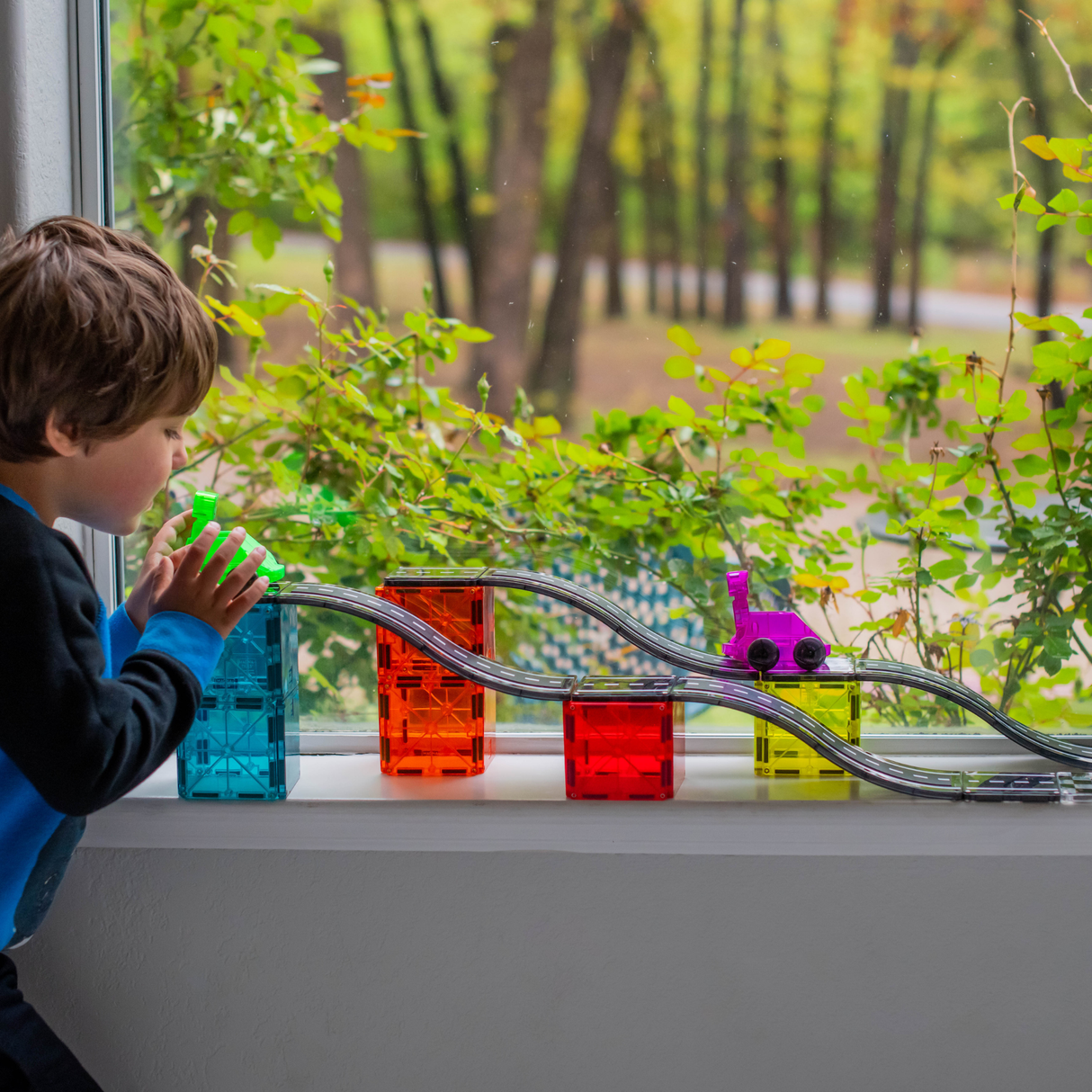 Child playing with colorful MAGNA-TILES® Downhill Duo 40pc set featuring ramps and vehicles on a windowsill track.
