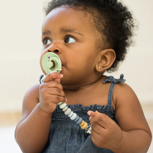 Baby holding a green pacifier attached to the Soft Multi Pacifier Clip with silicone beads and stainless steel clip.