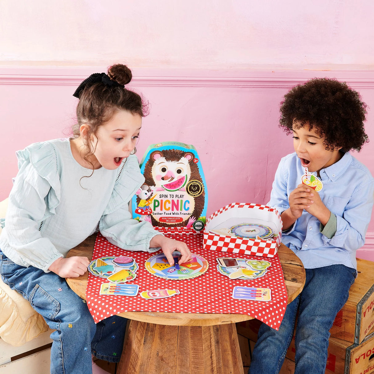 Two children playing the Picnic Spin to Play game with spinner and colorful picnic food pieces on a red tablecloth.