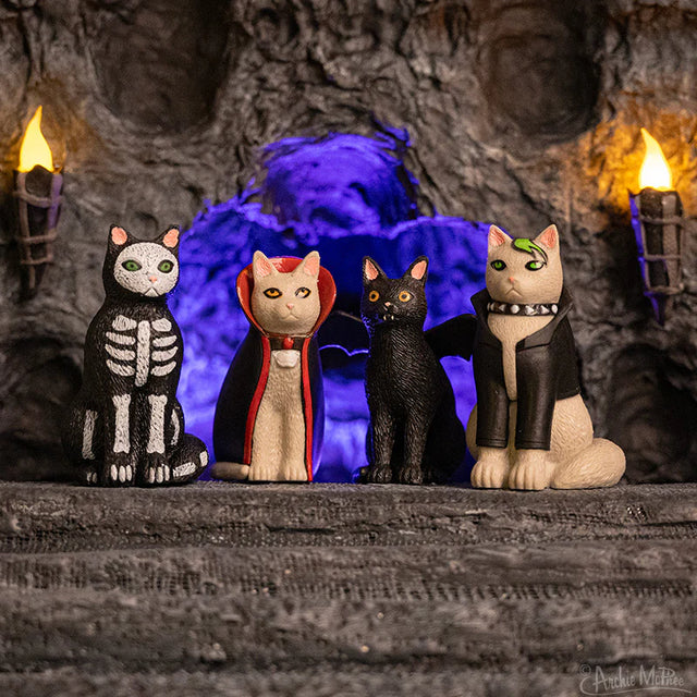 Four soft vinyl Goth Cat figurines in spooky costumes sitting against a cave backdrop with glowing blue light.