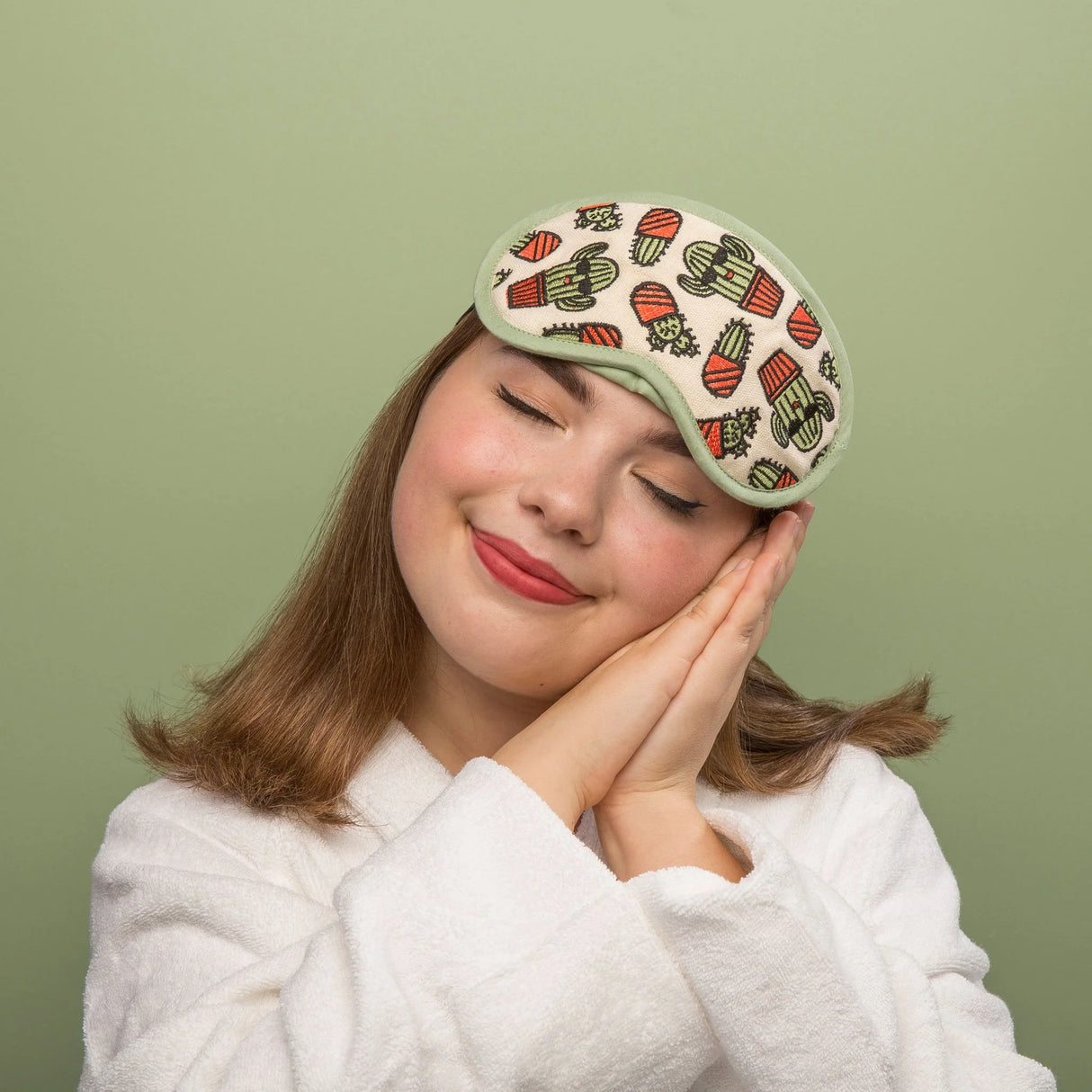 Young woman wearing a Happy Cactus Good Sleep Eye Mask with cactus print, resting peacefully with hands near face.