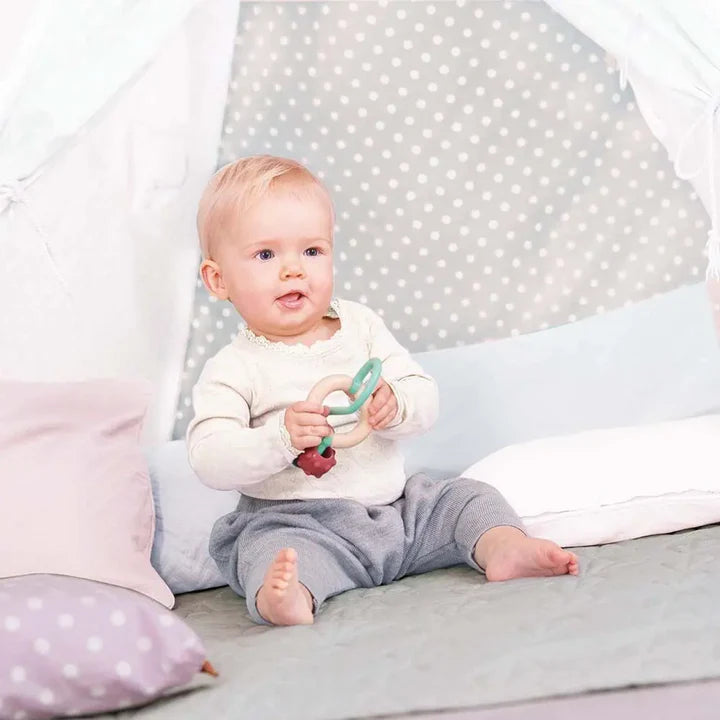 Baby sitting on soft cushions holding a Bead Maze toy with textured rings for sensory play and fine motor skills.