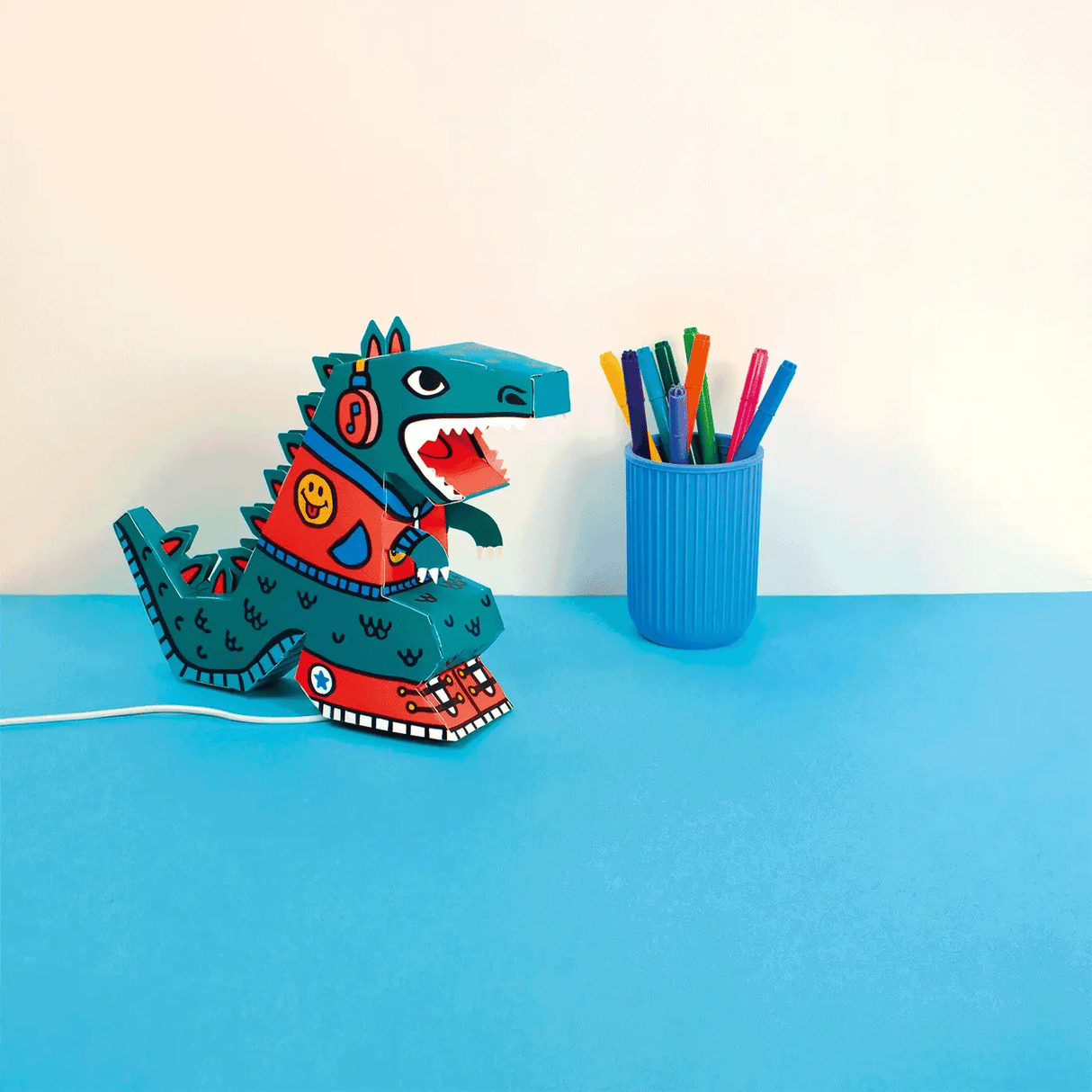 3D DIY Lamp | Dino assembled on blue surface near cup of colorful markers with USB cable plugged in.