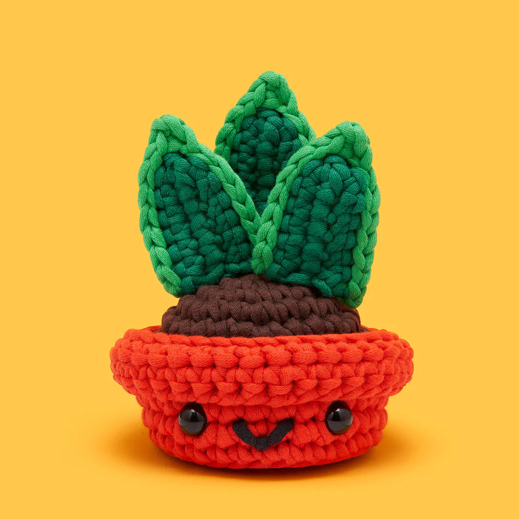 Crocheted snake plant with green leaves and a smiling face in a bright red pot against a yellow background